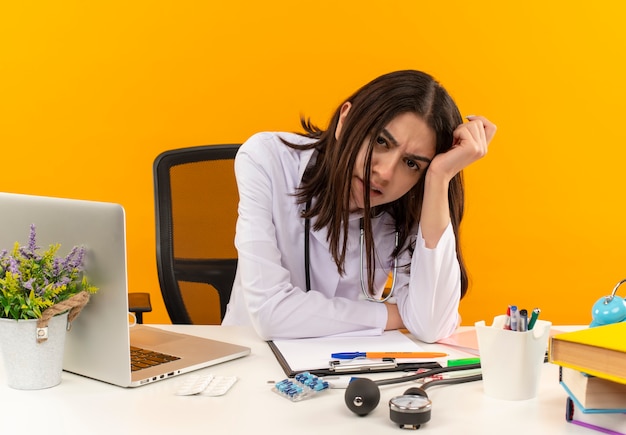 Joven doctora en bata blanca con estetoscopio mirando al frente confundida y muy ansiosa sentada en la mesa con computadora portátil y documentos sobre pared naranja