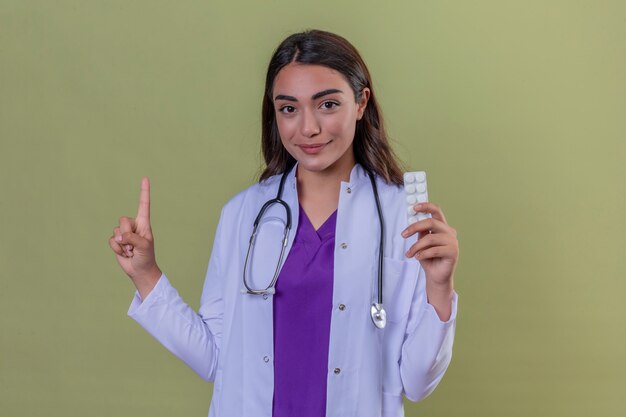 Joven doctora en bata blanca con fonendo sosteniendo blister con pastillas y apuntando hacia arriba con el dedo positivo y feliz de pie sobre fondo verde aislado