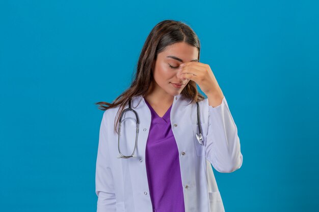 Joven doctora en bata blanca con fonendoscopio mirando cansado tocar la cabeza con la mano de pie sobre fondo azul aislado