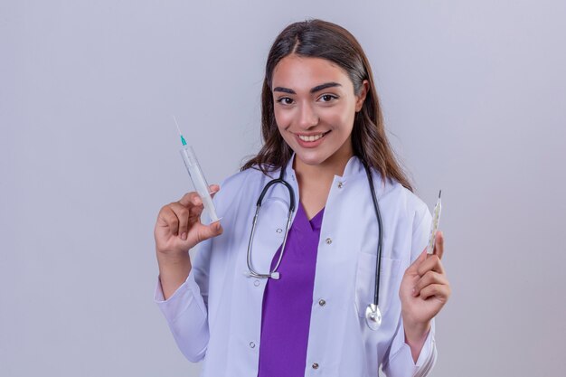 Joven doctora en bata blanca con fonendoscopio mirando confiado con una sonrisa en la cara con jeringa y termómetro sobre fondo blanco aislado