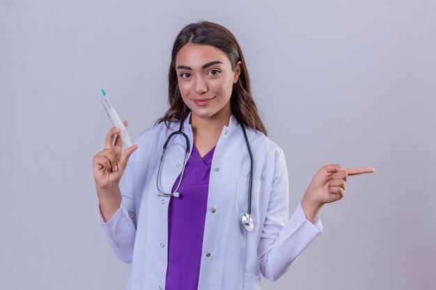 Joven doctora en bata blanca con fonendoscopio mirando confiado sosteniendo la jeringa con vacuna médica y apuntando con la mano y el dedo al lado sobre fondo blanco aislado