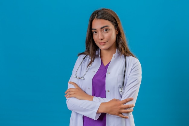 Joven doctora en bata blanca con fonendoscopio mirando seguro de pie con los brazos cruzados sobre fondo azul aislado