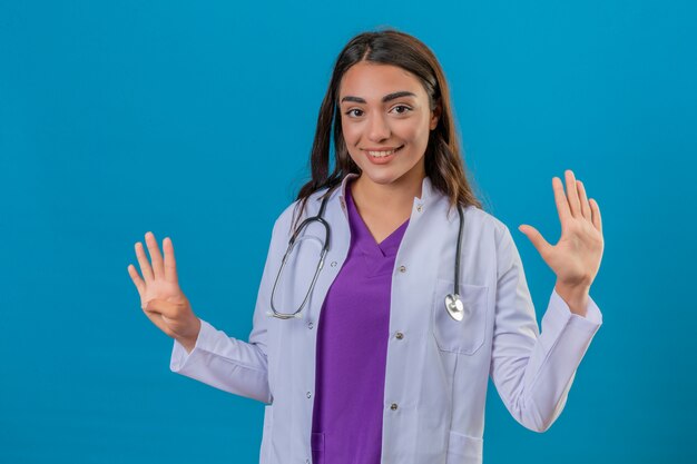 Joven doctora en bata blanca con fonendoscopio mostrando y apuntando hacia arriba con los dedos número nueve mientras sonríe seguro y feliz de pie sobre fondo azul aislado
