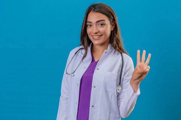 Joven doctora en bata blanca con fonendoscopio mostrando y apuntando hacia arriba con los dedos número tres mientras sonríe seguro y feliz de pie sobre fondo azul aislado