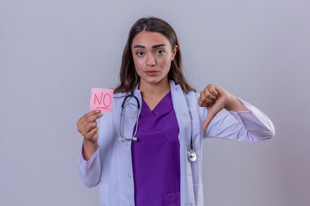 Joven doctora en bata blanca con fonendoscopio con papel recordatorio sin mensaje mostrando disgusto con los pulgares hacia abajo sobre fondo blanco aislado