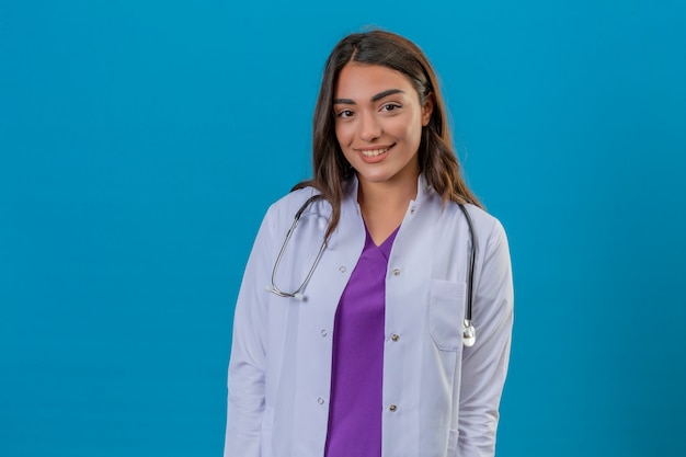 Joven doctora en bata blanca con fonendoscopio de pie con una gran sonrisa mirando a la cámara sobre fondo azul aislado