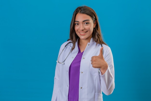 Joven doctora en bata blanca con fonendoscopio de pie con una gran sonrisa pulgar arriba sobre fondo azul aislado