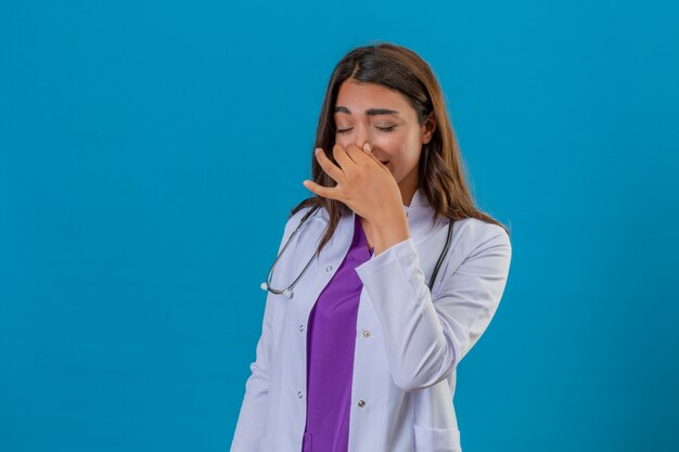 Joven doctora en bata blanca con fonendoscopio de pie con los ojos cerrados, conteniendo la respiración con los dedos en la nariz concepto de mal olor sobre fondo azul aislado