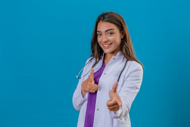 Joven doctora en bata blanca con fonendoscopio sonriendo y apuntando a la cámara con los dedos sobre fondo azul aislado