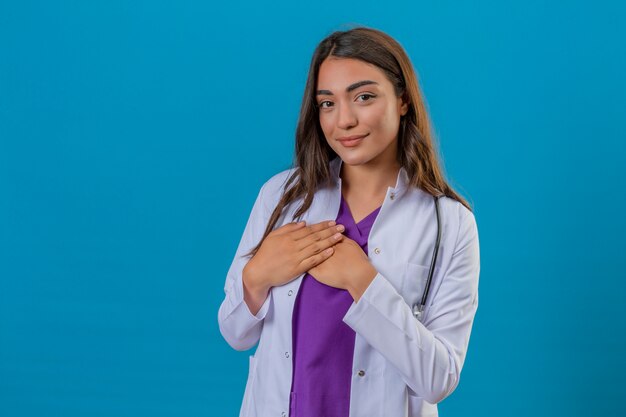 Joven doctora en bata blanca con fonendoscopio sonriendo con las manos en el pecho y agradecido gesto en la cara sobre fondo azul aislado