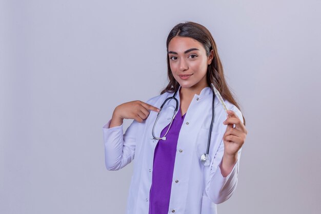 Joven doctora en bata blanca con fonendoscopio sonriendo sosteniendo el termómetro y apuntando con el dedo a sí misma sobre fondo blanco aislado