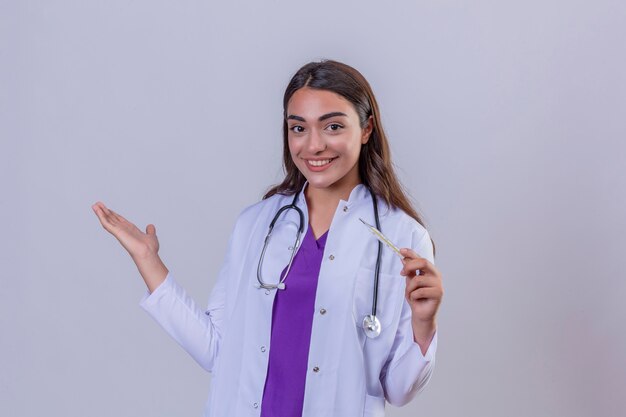 Joven doctora en bata blanca con fonendoscopio sonriendo sosteniendo el termómetro y con el brazo en un gesto de bienvenida sobre fondo blanco aislado