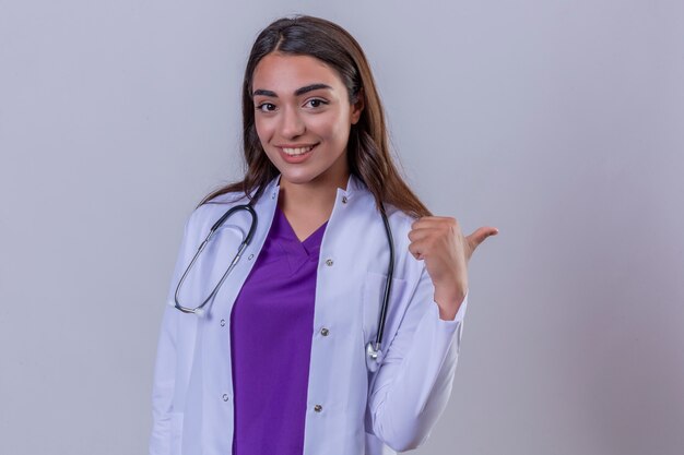 Joven doctora en bata blanca con fonendoscopio con sonrisa en la cara y apuntando hacia el lado con el dedo parado sobre fondo blanco.