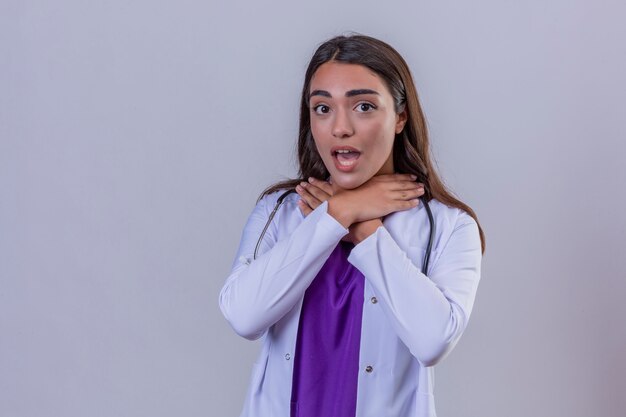 Joven doctora en bata blanca con fonendoscopio tocando su cuello y sufriendo de dolor de garganta sobre fondo blanco.