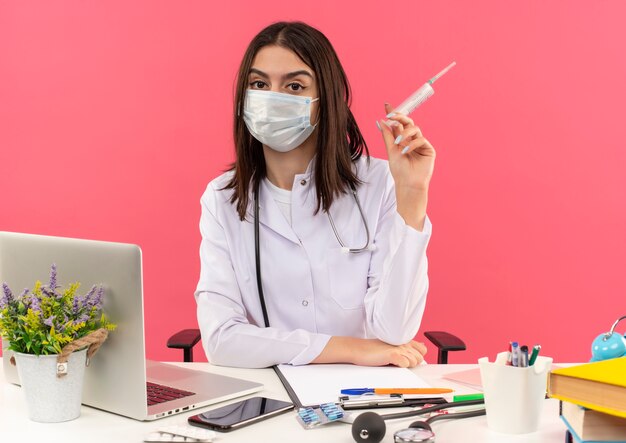 Joven doctora en bata blanca con máscara protectora facial y estetoscopio alrededor de su cuello sosteniendo una jeringa sentada en la mesa con un portátil sobre una pared rosa