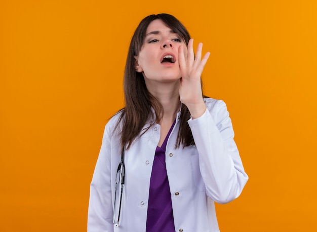 Joven doctora en bata médica con estetoscopio llama a alguien en la pared naranja aislada