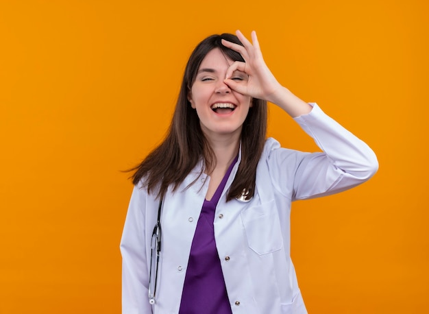 Joven doctora en bata médica con estetoscopio mira los dedos en la pared naranja aislada