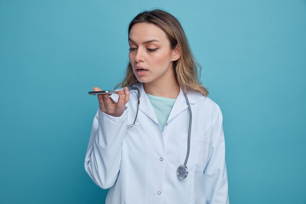 Joven doctora concentrada vistiendo bata médica y un estetoscopio alrededor del cuello sosteniendo y mirando el teléfono móvil hablando por su micrófono