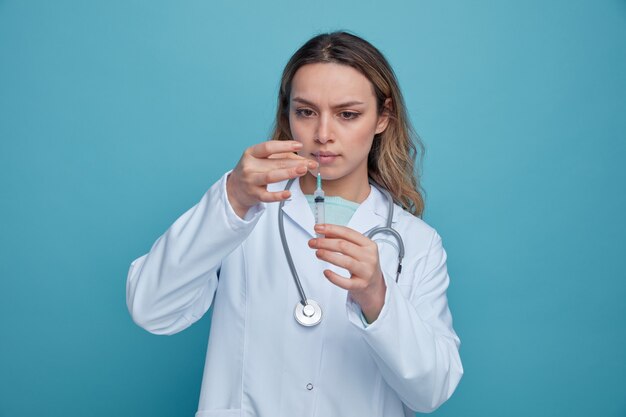 Joven doctora concentrada vistiendo una bata médica y un estetoscopio alrededor del cuello tocando la jeringa quitando las burbujas de aire