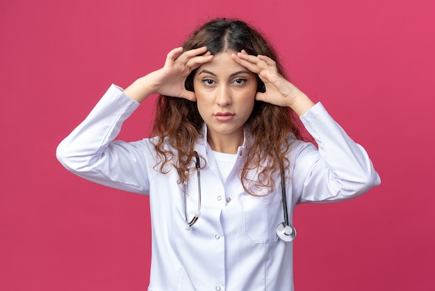 Joven doctora concentrada vistiendo bata médica y estetoscopio manteniendo las manos en la frente mirando al frente aislado en la pared rosa