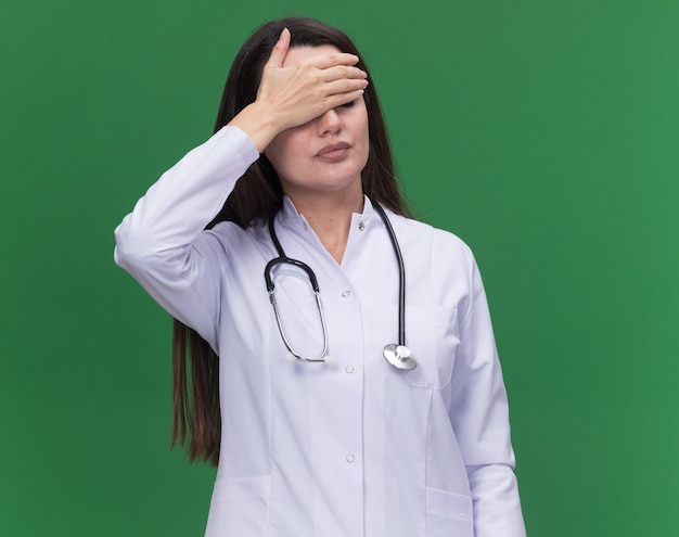 Joven doctora decepcionada vistiendo bata médica con estetoscopio pone la mano en la frente aislada en la pared verde con espacio de copia