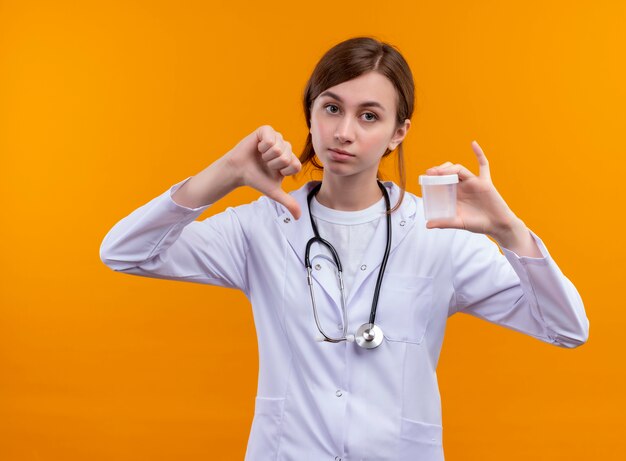 Joven doctora disgustada con bata médica y estetoscopio sosteniendo un vaso de precipitados y mostrando el pulgar hacia abajo en la pared naranja aislada