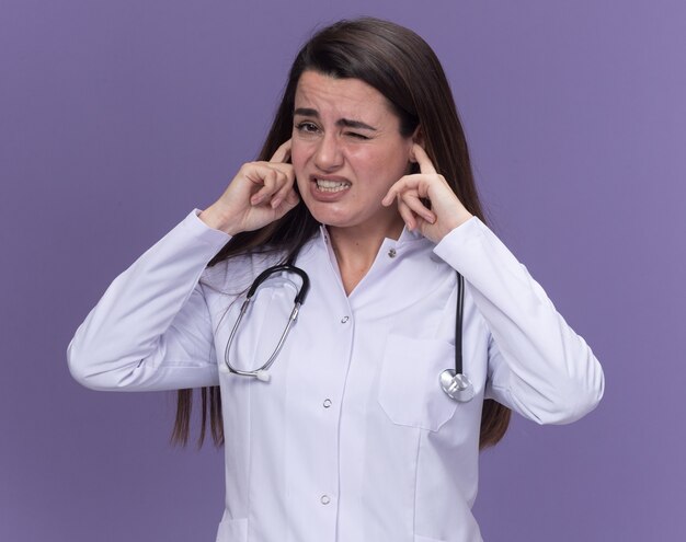 Joven doctora disgustada vistiendo bata médica con estetoscopio cierra las orejas con los dedos en púrpura