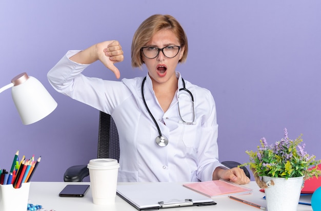 Joven doctora disgustada vistiendo bata médica con estetoscopio y gafas se sienta a la mesa con herramientas médicas que muestran el pulgar hacia abajo aislado sobre fondo azul.