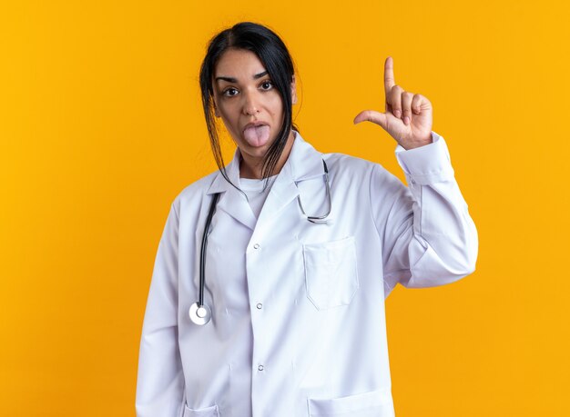 Joven doctora disgustada vistiendo bata médica con estetoscopio mostrando un gesto más suelto con la lengua aislada sobre fondo amarillo