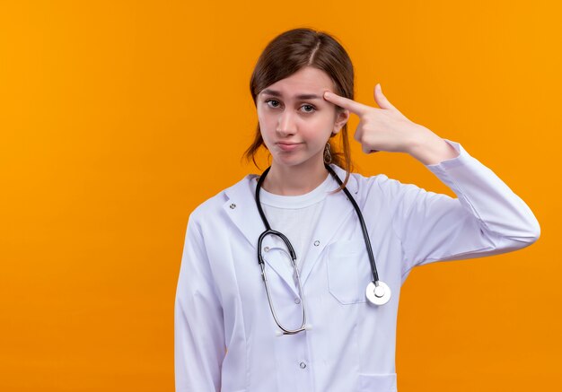 Joven doctora disgustada vistiendo bata médica y estetoscopio poniendo el dedo en la sien en la pared naranja aislada con espacio de copia