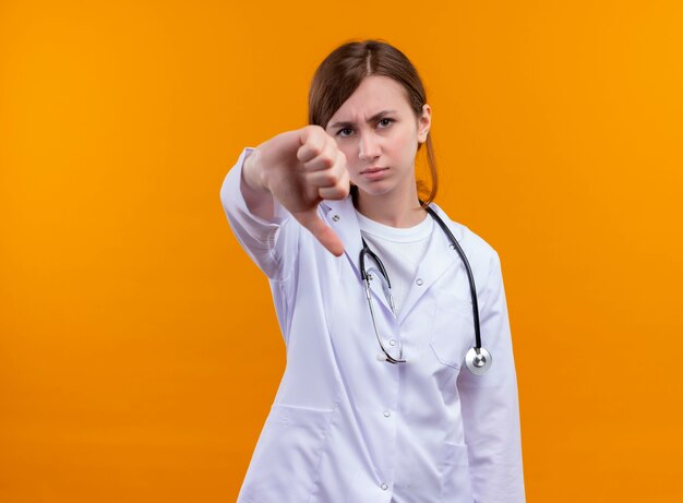 Joven doctora insatisfecha con bata médica y estetoscopio mostrando el pulgar hacia abajo en la pared naranja aislada con espacio de copia