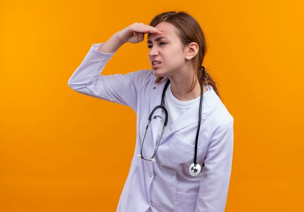 Joven doctora insatisfecha con bata médica y estetoscopio poniendo la mano cerca de la frente y mirando a distancia en la pared naranja aislada