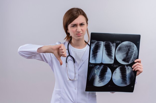 Joven doctora insatisfecha con bata médica y estetoscopio y sosteniendo una radiografía que muestra el pulgar hacia abajo en la pared blanca aislada