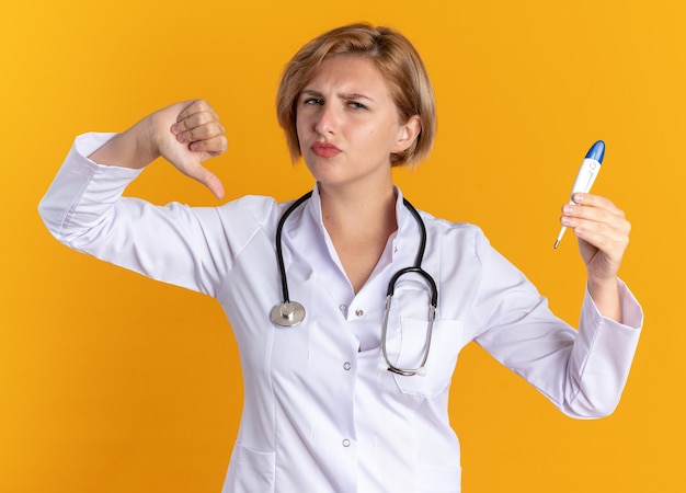 Joven doctora insatisfecha vistiendo bata médica con estetoscopio sosteniendo el termómetro mostrando el pulgar hacia abajo aislado sobre fondo naranja