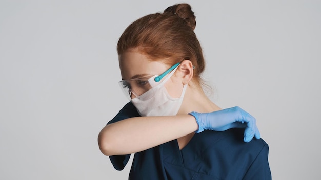 Joven doctora con máscara protectora para anteojos y guantes médicos tosiendo cubriendo con el hombro sobre fondo blanco.