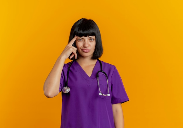 Joven doctora morena disgustada en uniforme con estetoscopio pone el dedo en la sien