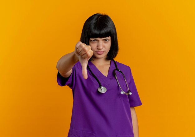 Joven doctora morena disgustada en uniforme con estetoscopio pulgar hacia abajo aislado sobre fondo naranja con espacio de copia