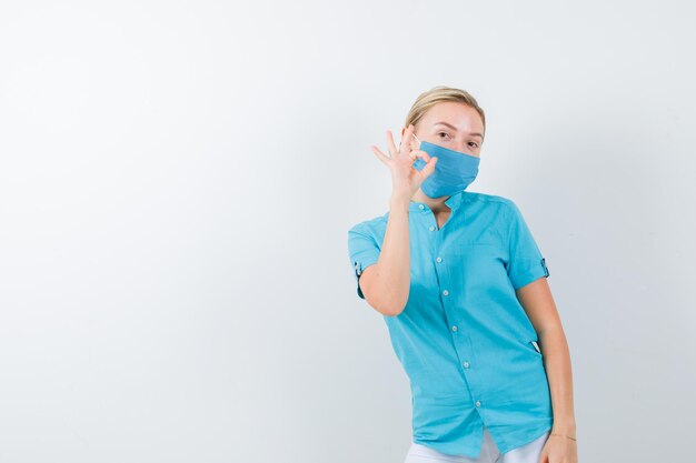 Joven doctora mostrando gesto ok en uniforme médico, máscara y mirando alegre