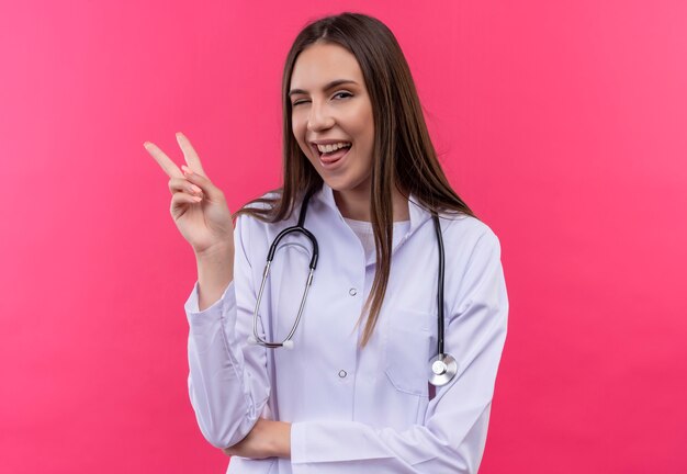 Joven doctora parpadeó con estetoscopio bata médica mostrando la lengua y el gesto de paz en la pared rosa aislada