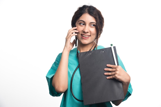 Joven doctora con portapapeles y hablando por teléfono celular sobre fondo blanco.