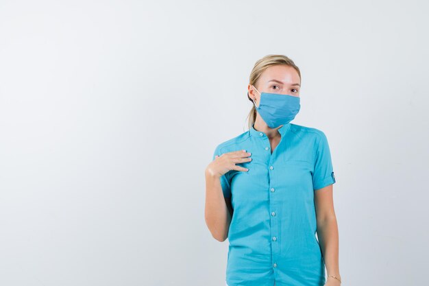 Joven doctora sosteniendo la mano en el pecho en uniforme y mirando indeciso