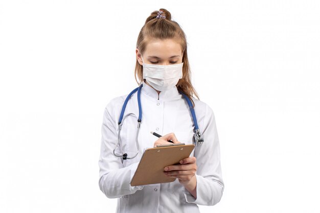 Joven doctora en traje médico blanco con estetoscopio en máscara protectora blanca escribiendo notas sobre el blanco