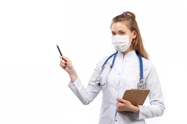 Joven doctora en traje médico blanco con estetoscopio en máscara protectora blanca escribiendo notas sobre el blanco