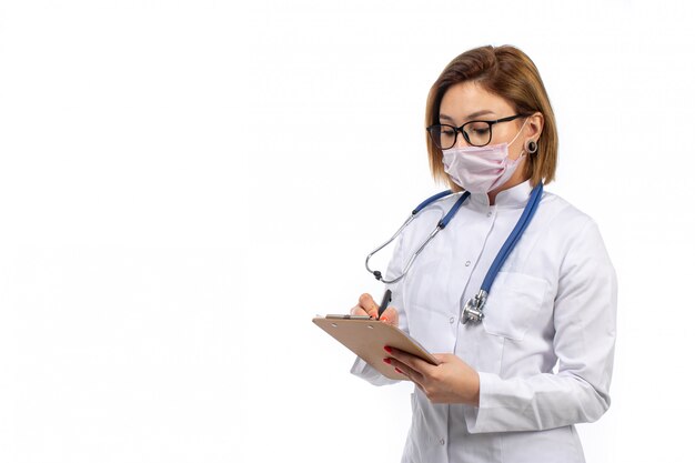 Joven doctora en traje médico blanco con estetoscopio en máscara protectora blanca escribiendo notas sobre el blanco