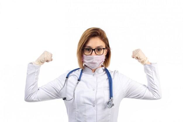 Joven doctora en traje médico blanco con estetoscopio en máscara protectora blanca flexionando sobre el blanco