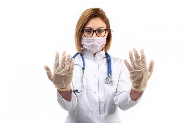Joven doctora en traje médico blanco con estetoscopio en máscara protectora blanca mostrando guantes en el blanco