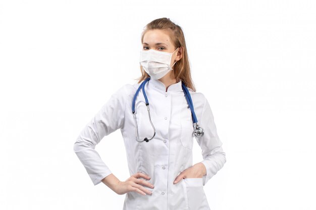 Joven doctora en traje médico blanco en estetoscopio de máscara protectora blanca posando en el blanco