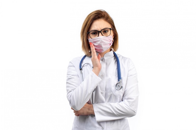 Joven doctora en traje médico blanco con estetoscopio en máscara protectora blanca sobre el blanco
