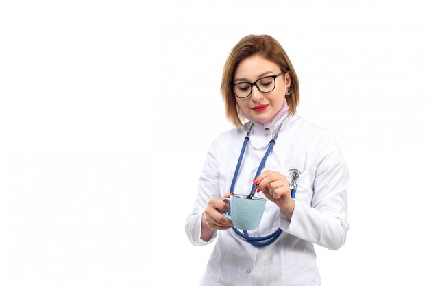Joven doctora en traje médico blanco con estetoscopio sosteniendo la taza azul sobre el blanco