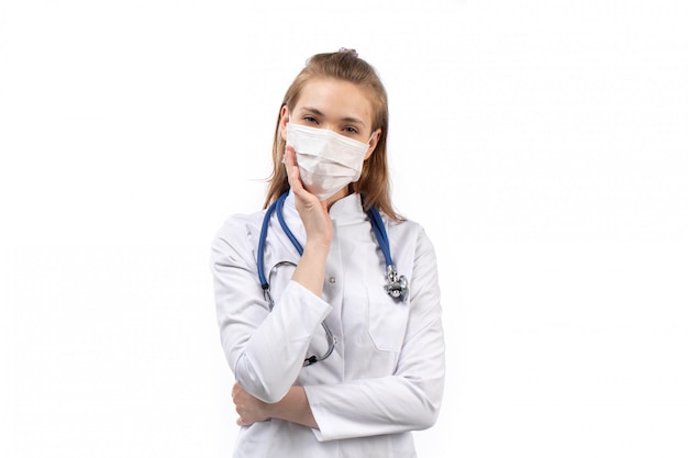 Joven doctora en traje médico blanco en máscara protectora blanca estetoscopio pensando pose en el blanco